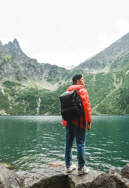 Vertical. Jovem turista de pé com as costas na margem de um belo lago na chuva em um fundo de montanhas, vestindo uma capa de chuva vermelha e um cap.Hiker fica em pedra perto do Lago Morskie Oko — Fotografia de Stock