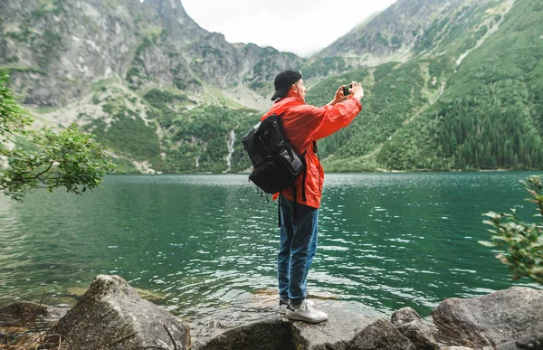 Young man in red raincoat and with backpack makes photo of lake in mountains on smartphone. Portrait of a tourist stands on the shore of Lake Morskie Oko and photographs the unreal scenery.