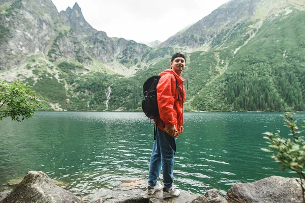 Portrait of happy young male tourist in red raincoat stands on background of lake and green mountains with coniferous trees, looks into camera and smiles. Morskie Oko, Tatra Mountains — 스톡 사진