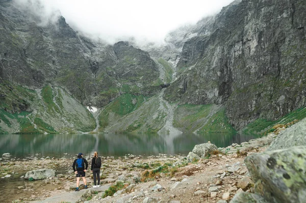 Para turystów stojąca na skałach i patrząca na górskie jezioro. Mężczyzna i kobieta z plecakami stoją plecami do przejrzystego jeziora i gór z chmurami. Tatry Zachodnie — Zdjęcie stockowe