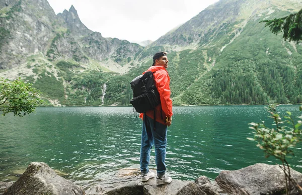 Full length portrait of a young tourist in a red raincoat and with a backpack stands against a background of a clear lake in the mountains and enjoys the mountains. Young man hiking in the rain. — 스톡 사진