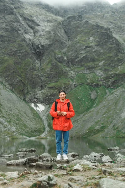 Retrato vertical de cuerpo entero de un joven senderista divertido con una chaqueta roja con un smartphone en la mano sobre el fondo de montañas y lagos, mira a la cámara y sonríe. Hombre y naturaleza — Foto de Stock