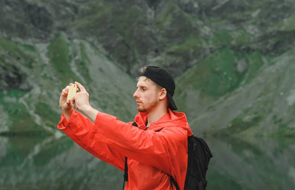 Handsome young tourist in mountains near lake makes nature photo on smartphone. Portrait of a tourist on a mountain hike taking a selfie. Guy in the red jacket photographs the mountain scenery.