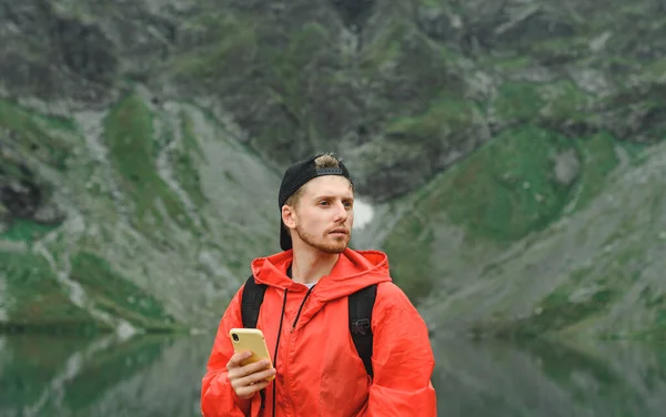 Retrato de close-up de um jovem com uma capa de chuva vermelha fica com um smartphone na mão em um fundo de montanhas, olhando para longe. Guy turista em um chapéu fica nas montanhas e usa a Internet . — Fotografia de Stock
