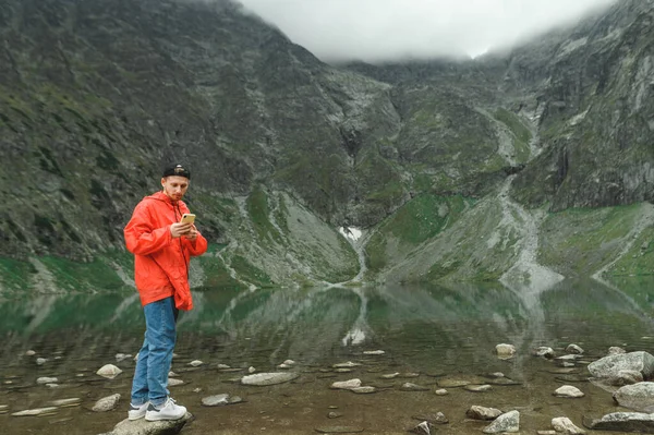 Young male tourist in red jacket stands on a rock in the mountains and uses a smartphone against a background of a mountain lake.Full length portrait of hiker on hike standing with smartphone in hand — 스톡 사진