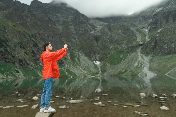 Bonito jovem turista em montanhas perto do lago faz foto no smartphone.Portrait de um turista em uma caminhada na montanha tomando uma selfie.Guy na jaqueta vermelha fotografa a paisagem da montanha com o lago — Fotografia de Stock