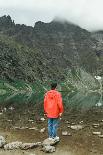 Retrato de comprimento total de um jovem caminhando, em pé no topo de uma montanha perto de um lago e olhando para a bela paisagem. Foto vertical de turista em jaqueta vermelha em montanhas perto do lago. Espaço de cópia — Fotografia de Stock