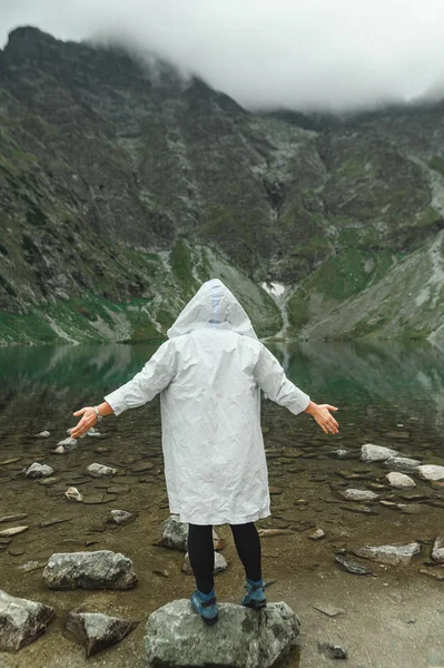 Pessoa em uma capa de chuva branca fica em uma rocha perto de um lago de montanha e levanta as mãos e goza da cenária.Foto de turista com rotação no fundo do lago bonito irreal no topo da montanha — Fotografia de Stock