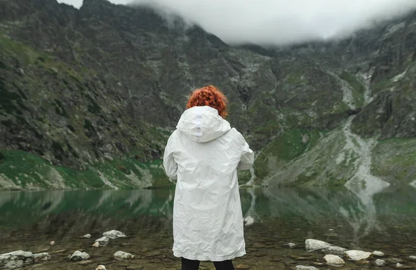 Woman with curly red hair and in a white cloak stands on the lake on a background of mountains, back view. Man and untouched nature.Hike to mountain lake concept. Background. Copy space