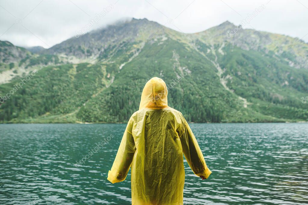 Background. A person in a yellow raincoat stands in the rain against the backdrop of a beautiful landscape with a lake and mountains, looking out at the view. Background. Hiking mountains concept.