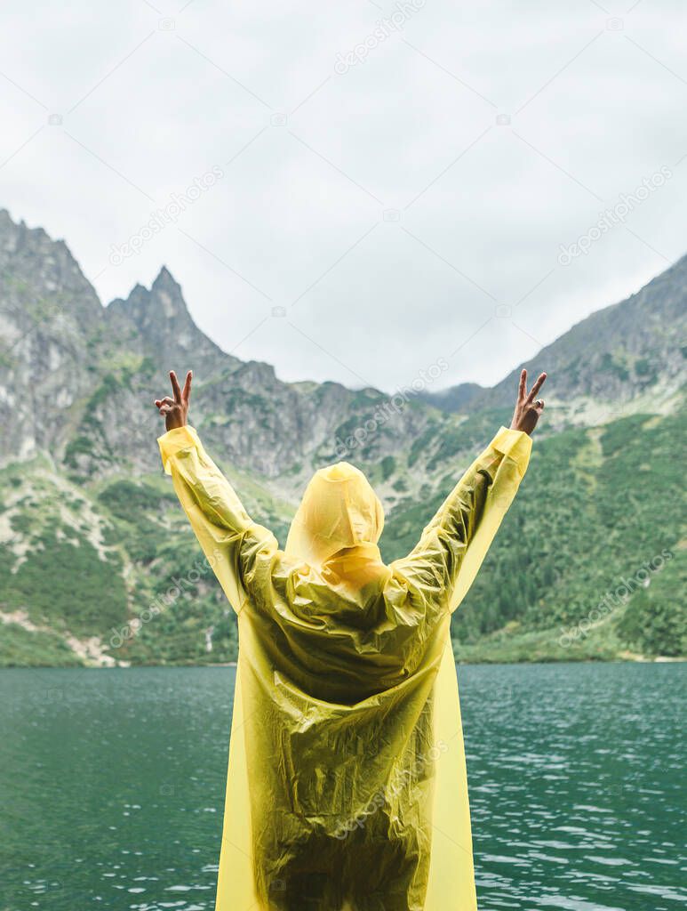 Vertical. Background. Copy space. Person in a yellow raincoat with his hands raised stands in the rain against the backdrop of a mountain lake and a mountain.Hiker hiking on Lake Morskie Oko