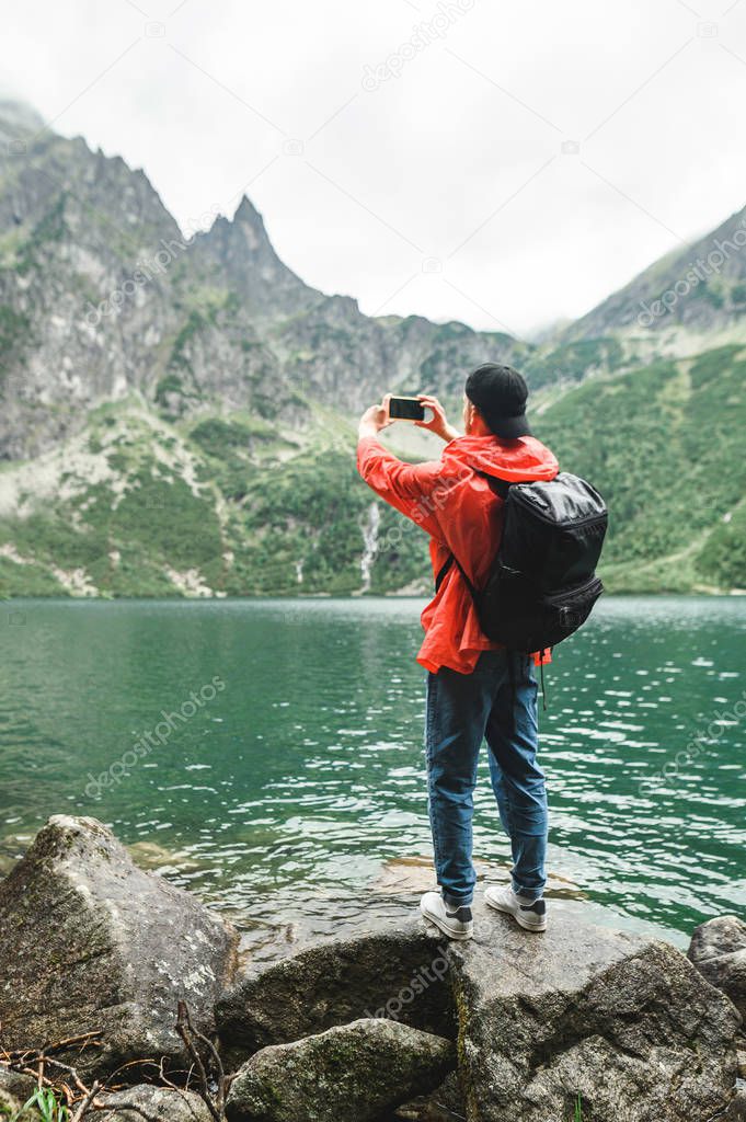 Hiker taking pictures of landscape. A young man with a backpack and a red raincoat stands on a rock on the shore and takes photo of a mountain lake and mountains on a smartphone camera.Vertical photo