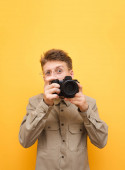 Portrét legračního fotografa v košili a brýlích pořídí fotografii na kameře na žlutém pozadí a podívá se do kamery s legračním obličejem. Legrační paparazzi fotí na kameru. Izolované.