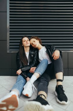 Karanlık bir arka planda oturan, koyu renk giyinen, kameraya sarılan ve poz veren iki şık kızın dikey fotoğrafı. Koyu renk arka planda deri ceketli iki mankenin portresi.