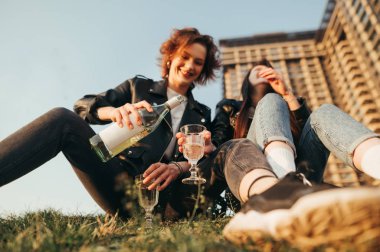 Gün batımında çimenlerde oturan ve bardaktan şarap dolduran gülümseyen kızlar. Yürüyüşte içki içen mutlu kız arkadaşlar. Neşeli bir kızın elindeki şişe şarap ve bardağa odaklan. Arkaplan.