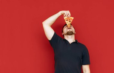 Siyah tişörtlü komik bir adam kırmızı arka planda pizza yiyor, kafasını kaldırıp ağzına bir parça sokuyor. Son dilim pizzayı yiyen adamın fotoğrafı, izole edilmiş. Fast food. Boşluğu kopyala