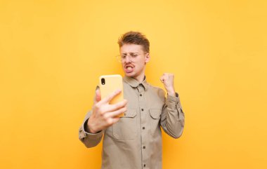 Gözlüklü, bıyıklı ve tişörtlü neşeli inek telefon ile sarı arka planda duruyor, ekrana bakıyor ve elini kaldırarak seviniyor. Akıllı telefonlu komik inek kazandı, sevindi..