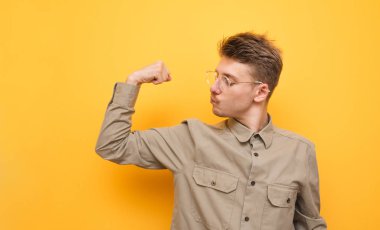 Bıyıklı ve gözlüklü komik bir genç adam gömlek giyiyor, sarı arka planda duruyor ve pazılarını gösteriyor. Gömlekli komik inek pazılarda kaslarını gösteriyor. İzole edilmiş.