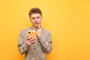 Gözlüklü ve gömlekli dalgın genç adam elinde telefon, sarı arka planda duruyor, başka tarafa bakıyor ve düşünüyor. Düşünceli inek telefonunu kullanır ve fotokopi odasına bakar.