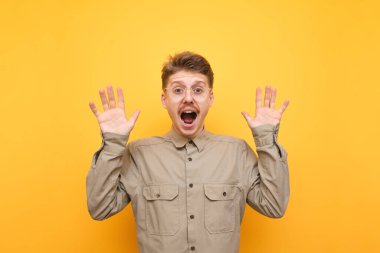 Sarı arka planda, korkmuş gözlüklü ve gömlekli genç adamın portresi, korku içinde ellerini kaldırdı ve şok olmuş yüzüyle kameraya baktı. Korkak inek çocuk şok olmuş suratıyla kameraya bakıyor..