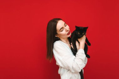 Beyaz gömlekli neşeli kadın kırmızı arka planda siyah kediyle oynuyor, el ele tutuşuyor, evcil hayvana bakıyor ve gülümsüyor. İzole edilmiş. Uzayı kopyala Evcil hayvan konsepti.
