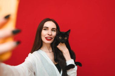 Beyaz gömlekli çekici bir kadın kırmızı arka planda siyah kediyle selfie çekiyor, akıllı telefon kamerasına bakıyor ve gülümsüyor. Ev yapımı ovalama ile selfie. Kırmızı arkaplanda izole.