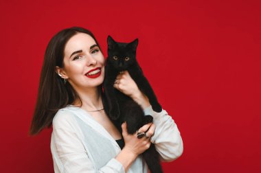 Kırmızı arka planda sevimli siyah bir kediyle sarılıp kameraya bakıp gülümseyen neşeli bir kızın portresi. Beyaz gömlekli mutlu bayan evcil bir hayvana sarılıyor. İzole edilmiş.