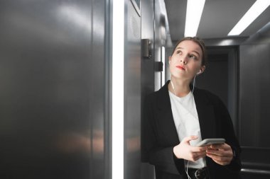 Asansörde kat numaradan arıyorum ve onu akıllı telefon ve kulaklık kullanarak iddialı ofis çalışanı portresi. Genç işkadını ararken telefon ve kulaklık ile asansörde.