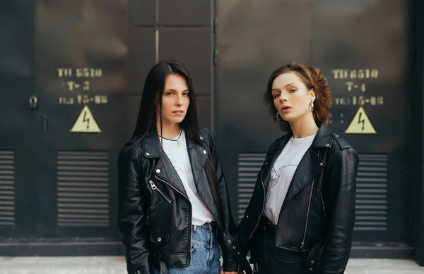 2 beautiful models standing against a dark wall background and posing at the camera, Two girls in a leather jacket stand on the black wall background and looking at the camera with a serious face. — 스톡 사진
