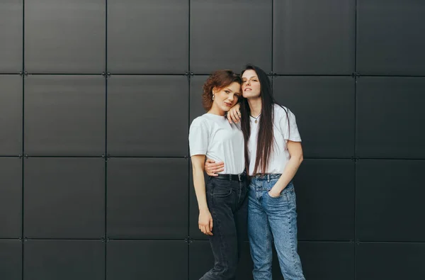 Attractive girls in white T-shirts hugging on a black background, looking at the camera and smiling. Couple of happy girlfriends are standing on black background and posing at camera — 스톡 사진