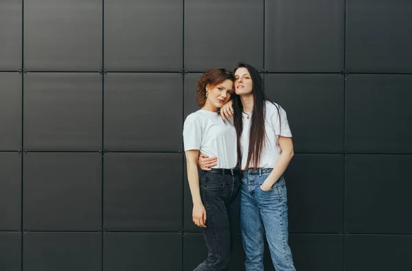 Meninas atraentes em camisetas brancas abraçando um fundo preto, olhando para a câmera e sorrindo. Casal de namoradas felizes estão de pé no fundo preto e posando para a câmera — Fotografia de Stock