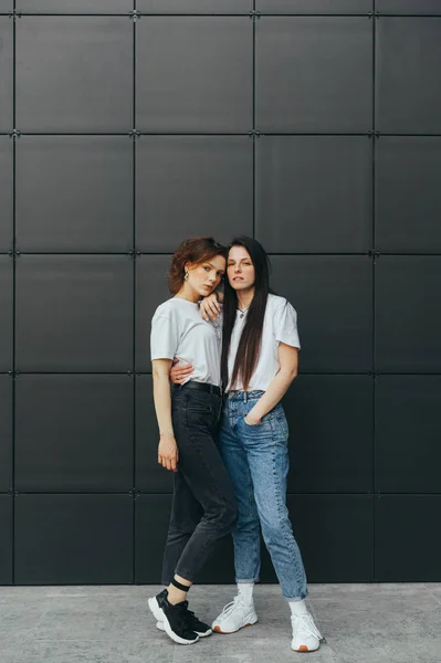 Foto vertical. Meninas bonitas em baixas abraçando no fundo preto e olhando para a câmera. Casal de belas namoradas estão de pé sobre fundo preto e posando para a câmera. Espaço de cópia — Fotografia de Stock