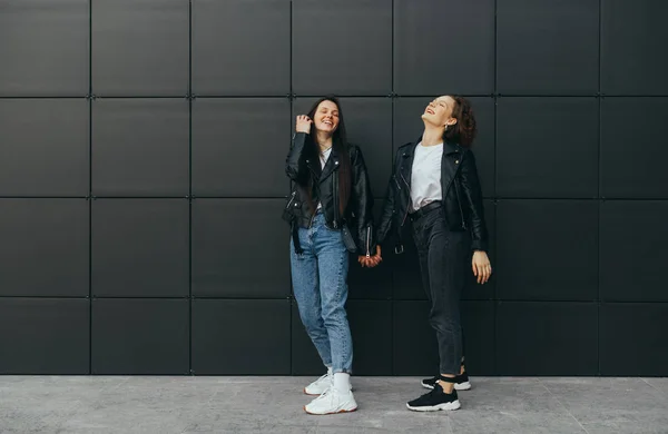 Two girlfriends having fun on dark background. Couple of happy girls in stylish street clothes are standing against a black wall background,holding hands and smiling. Urban photo of two positive girls — Stock Photo, Image