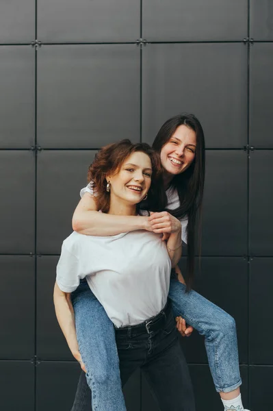Portrait of cheerful two girlfriends having fun on dark background, girl carry girlfriend on her back, piggy back, looking at camera and smiling. Happy 2 girls having fun on black wall background — 스톡 사진