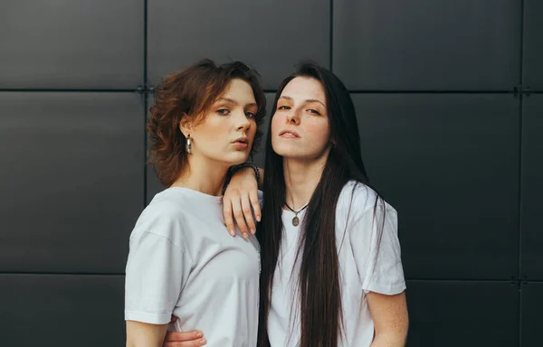 Retrato de close-up de duas meninas atraentes em camisetas brancas em pé contra um fundo de parede preta e posando para a câmera. Foto na moda de um par de meninas em um fundo escuro. Espaço de cópia — Fotografia de Stock