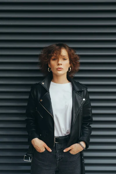 Fashionable photo curly-haired girl in casual clothes stands against a dark background and looks into the camera, serious face wearing a leather jacket.Girl with short hair on a black wall background — 스톡 사진