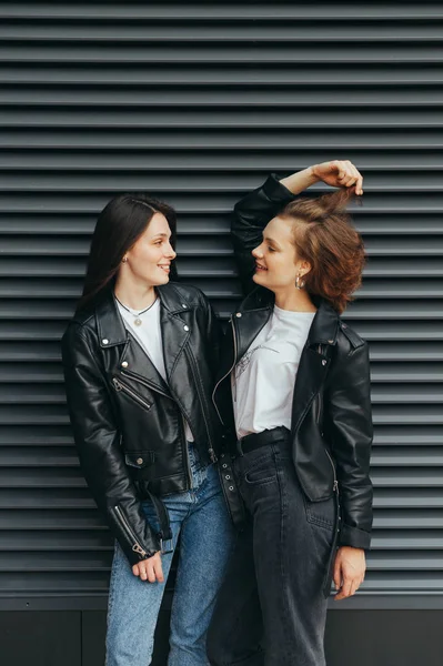Щаслива пара дівчат у повсякденному одязі стоїть на фоні чорної стіни, розмовляє та усміхається, одягнена в шкіряні куртки. Дві веселі подружки сміються на темному тлі. Вертикальний — стокове фото