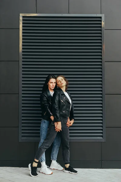 Two girls in dark casual clothes stand on a black background, hugging and posing at the camera.Attractive best girlfriends are standing against a dark wall and looking at camera with a serious face — 스톡 사진