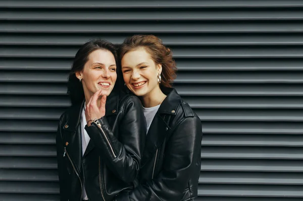 Retrato de close-up de um par de meninas felizes em fundo preto, falando e sorrindo, vestindo roupas casuais escuras. Sorrindo namoradas em jaquetas de couro ficar contra um fundo escuro e se divertir — Fotografia de Stock