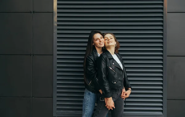 Two happy girlfriends in casual clothes hugging against a dark wall, looking at the camera and smiling, wearing leather jackets. 2 positive girls are standing outside on a dark background and hugging — 스톡 사진