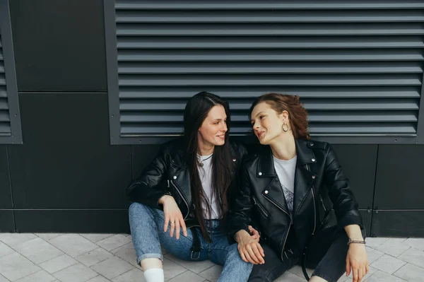 Close up photo of two smiling girls in leather jackets and jeans sitting on a cobblestone against a dark wall background and smiling.Girlfriends sat down for a break on the walk,looking at each other — 스톡 사진