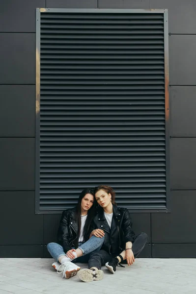 Duas meninas atraentes em roupas casuais sentam-se no chão contra uma parede escura, olhando para a câmera com um rosto sério, vestindo jaquetas de couro. 2 amigas sentam-se em um pavers e posam. Espaço de cópia . — Fotografia de Stock