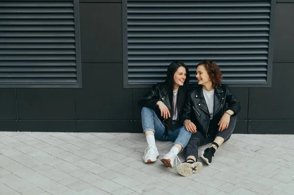 Duas meninas felizes em roupas casuais elegantes estão sentadas em um pavimento contra um fundo de parede escura, olhando um para o outro e sorrindo. Foto de rua de duas namoradas descansando, agachadas no chão — Fotografia de Stock