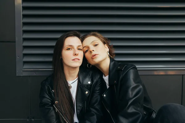 Closeup photo of two girls with freckles on a dark background, wearing dark clothes,posing for the camera. Portrait of 2 models in leather jackets on dark wall, looking in camera with serious face — Stock Photo, Image