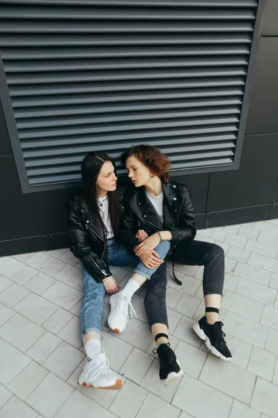 Modieuze foto van twee meisjes in leren jassen en jeans zittend op een ondergrond tegen een donkere muurachtergrond. Vriendinnen gingen zitten voor een pauze op de wandeling, meisje kijkt naar vriendin — Stockfoto