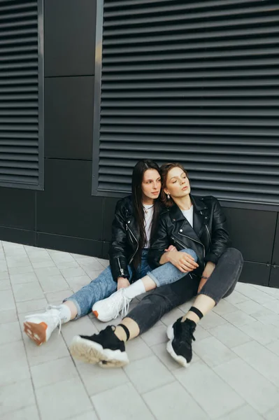 Módní fotografie 2 stylových dívek v neformálním oblečení, v kožené bundě, sedících na zemi u tmavé stěny a hledících pryč.Ulice portrét dvou přítelkyň sedících na chodníku.Vertikální — Stock fotografie