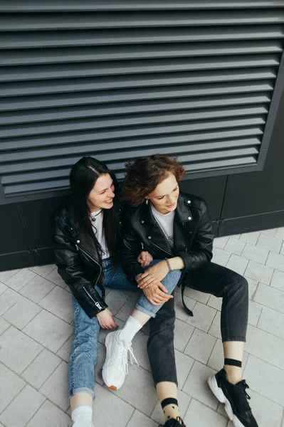 Due ragazze attraenti in eleganti vestiti casual seduti per strada a terra a parlare e sorridere. Foto di vista dall'alto alla moda di due amiche felici che ingannano per strada. Ricevuto. Verticale — Foto Stock