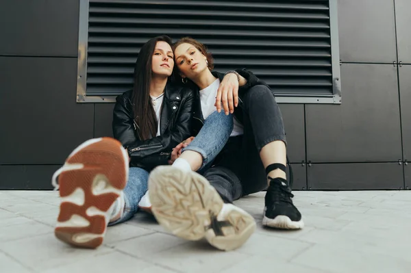 Photo à la mode de deux filles élégantes en vêtements décontractés portant des vestes en cuir et des baskets, assises sur le sol contre un mur sombre et posant en caméra.Portrait de mode 2 copines dans la rue — Photo