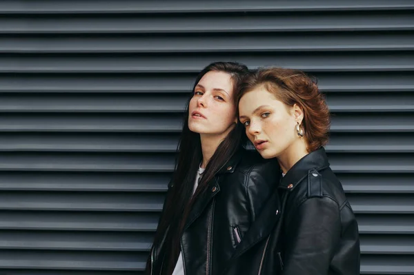 Retrato de duas meninas bonitas elegantes em roupas casuais em um fundo de parede preta, olhando para a câmera com um rosto sério. Dois modelos em jaquetas de couro posando para a câmera, fundo de parede escura — Fotografia de Stock