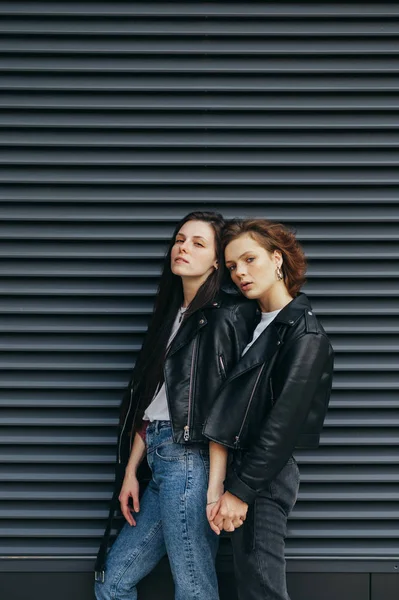 Dois modelos de roupas casuais elegantes ficam em um fundo preto, olhe para a câmera com um rosto sério. Duas meninas em jaquetas de couro posando na câmera contra o fundo da parede escura. Vertical — Fotografia de Stock